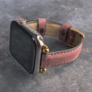 May include: A silver smartwatch with a black screen and a red ring on the side button. The watch features a dusty rose-colored leather strap with white stitching, secured by gold-toned metal hardware and a matching gold-toned buckle.