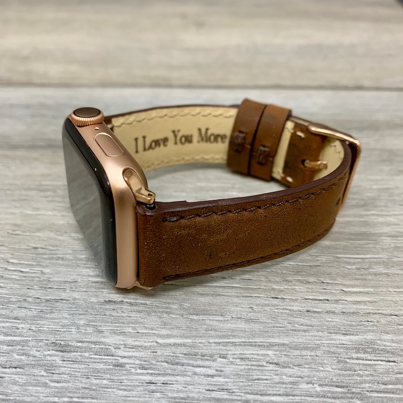 May include: Rose gold Apple Watch with a brown leather band. The band has a gold buckle and the words "I Love You More" are printed on the inside of the band. The watch is resting on a gray wooden surface.