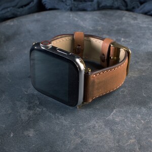 Puede incluir: Una correa de reloj de cuero marr&oacute;n con un Apple Watch plateado y negro.