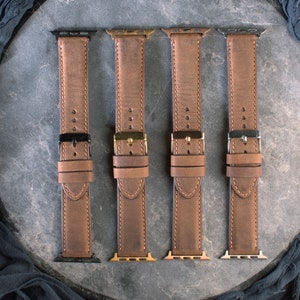 May include: Four brown leather watch straps with different coloured metal clasps. The straps are laid out on a grey surface.