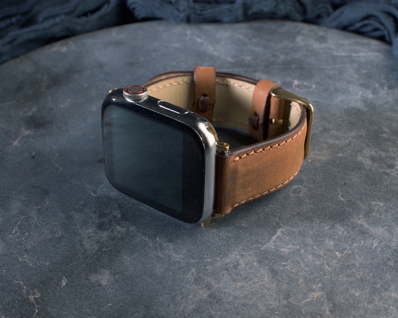 May include: A brown leather watch band with a stainless steel watch face. The band has a buckle closure and is made of a textured, distressed leather. The watch face has a black screen and a red button.