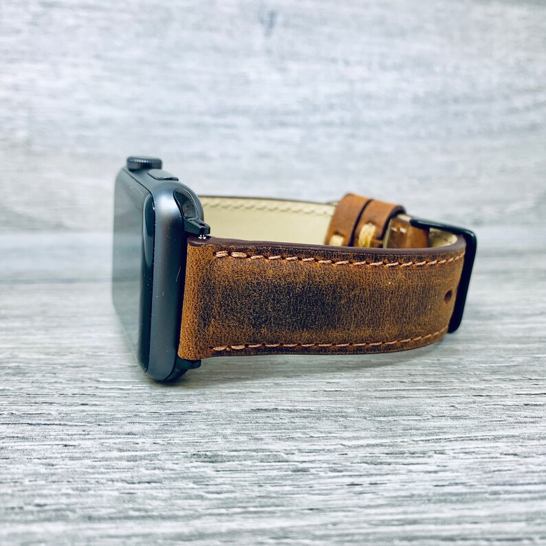 May include: A brown leather watch band with distressed detailing and a light brown stitching. The band is attached to a black Apple Watch.