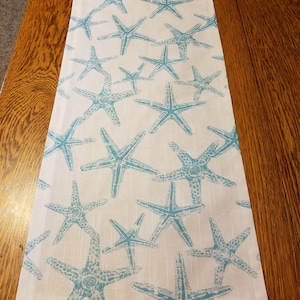 May include: A rectangular white table runner with a repeating pattern of turquoise starfish. The starfish vary in size and are evenly spaced across the fabric. The runner is made of a textured material.