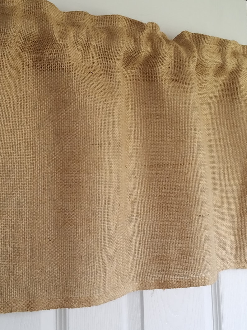 K&ouml;nnte beinhalten: Eine braune Jute-Gardine mit gerafftem Oberteil und geradem Unterteil. Die Gardine ist aus einem gewebten Stoff mit rauer Textur.