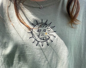 May include: Sage green t-shirt with an embroidered sun and moon design. The celestial design features a smiling sun and moon intertwined, with black and gold thread detailing. A silver Libra necklace is also visible.