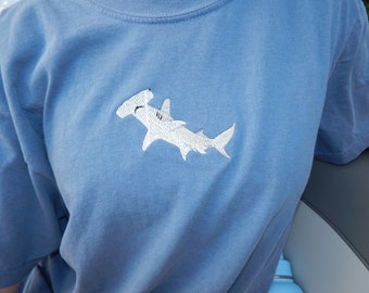 May include: A pale blue t-shirt with a white embroidered hammerhead shark motif. The top has a crew neck and short sleeves. The material looks soft and comfortable. The design is on the chest.