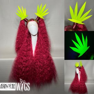 May include: A long, curly, red wig with a white lace front, styled with two neon green leaf-shaped hair accessories. The wig is displayed on a mannequin head. The text "GEORGINA THE DOLL WIGS" is visible.