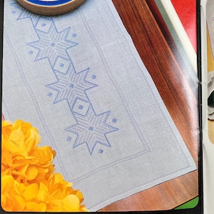 May include: An open craft book displaying two pages with embroidery projects. The left page shows a light blue table runner with a geometric star pattern. The right page features a lunch set with pink embroidered designs on light fabric.