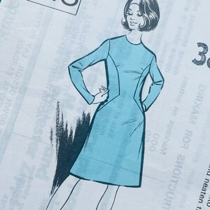 Peut inclure: Une pochette de patron de couture blanche marquée Pattern People No. 448, taille 38. Elle présente une illustration en noir et blanc d'une personne portant une robe bleu clair à manches longues. Le texte inclut des instructions et l'adresse.