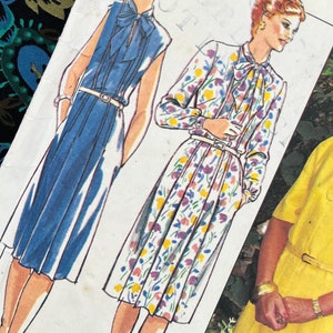 Peut inclure: Une pochette de patron de couture pour une robe avec col lavallière et ceinture. La couverture présente des illustrations d'une robe bleue unie et d'une robe à fleurs, ainsi qu'une photo d'une personne portant une robe jaune. Texte : Style et 12+14+16+18.