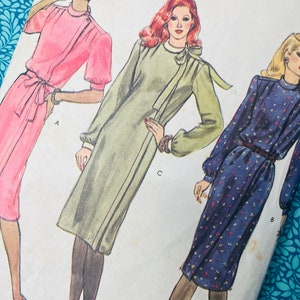 May include: A sewing pattern envelope featuring illustrations of three different dress styles. The first is a pink dress with a belted waist, the second is a sage green dress with a tie neck, and the third is a navy blue dress with a small floral print.