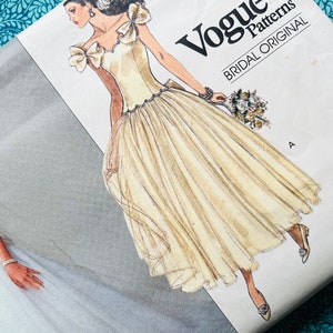 Może przedstawiać: Wzór szycia Vogue Patterns Bridal Original. Wzór przedstawia rysunek panny młodej w kremowej sukni ślubnej z pełną spódnicą i detalami kokardki. Koperta wzoru znajduje się na niebieskim tle kwiatowym.