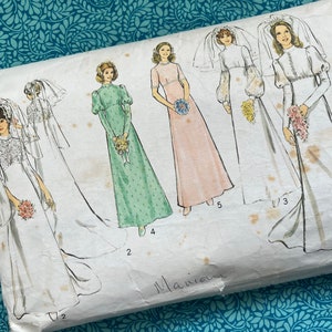 May include: Vintage sewing pattern featuring illustrations of wedding dresses. The pattern packet shows various bridal gown designs, including long-sleeved and sleeveless options, with veils and bouquets. The background is a teal floral pattern.