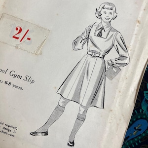 May include: A vintage paper sewing pattern envelope titled Practical Paper Pattern for a School Gym Slip in size 6-8 years. The cover features a black and white illustration of a person wearing a collared shirt, tie, and a pleated gym slip dress.