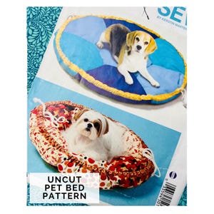 May include: An uncut pet bed pattern featuring two dog bed designs. One bed is a round, blue and yellow bed with a dog on it. The other bed is oval-shaped, with a red and white floral pattern, and a dog inside.