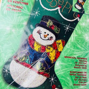 Peut inclure: Un kit de bas de Noël en appliqué de feutre avec un motif de bonhomme de neige fantaisiste. Le bas vert présente un bonhomme de neige portant un chapeau haut de forme et une écharpe colorée. Le kit comprend le texte "BONHOMMES DE NEIGE FANTAISIE Bas de Noël 18" (45,7cm) Créé par Bonnie Smith."