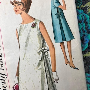 Op de afbeelding: Een vintage Simplicity naaipatroon-envelop met illustraties van een mouwloze shift-jurk met een strik aan de zijkant. De hoofdjurk is wit met een subtiel geometrisch patroon en een bloemaccent, terwijl de tweede variant effen blauw is.