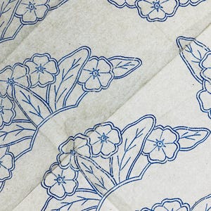 May include: A light gray paper surface featuring a repeating pattern of blue ink-stamped floral motifs. Each motif consists of five-petaled flowers and pointed leaves arranged in a fan-like cluster, with visible texture in the blue ink lines.