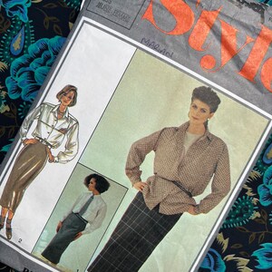 May include: A Style brand sewing pattern envelope numbered 4493, featuring illustrations of a long-sleeved button-down shirt and a long skirt. The cover displays the text Style, 12 MISS JEUNE FEMME, and handwritten names. The paper envelope is creased.