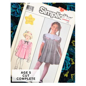 Könnte beinhalten: Ein Simplicity-Schnittmusterumschlag mit Illustrationen eines Kindes in einem rosa Mantel mit Hut und eines Kindes in einem schwarz-weiß karierten Kleid mit geschwungenem Kragen. Der Umschlag enthält den Text "made in heaven" und "AGE 5 CUT COMPLETE".