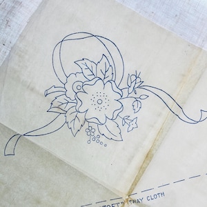 May include: Vintage floral embroidery pattern on translucent paper. The design features a large flower with a dotted center, leaves, and a ribbon, all in blue ink. The text "TOFT" TRAY CLOTH" is visible at the bottom.
