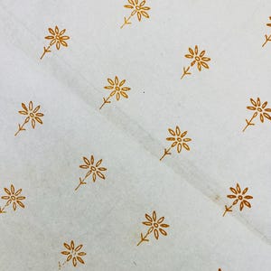 Könnte beinhalten: Ein Bogen cremefarbenes, halbtransparentes Papier mit einem sich wiederholenden Muster aus kleinen, senfgelben, gänseblümchenartigen Blumen. Jede Blume besteht aus einem zentralen Punkt, umgeben von dünnen Blütenblättern und einem kurzen Stiel.