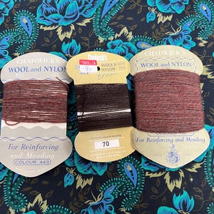 May include: Three spools of Chadwick's Wool and Nylon thread in shades of brown and black. The spools are labelled for reinforcing and mending, with colour numbers and thread composition details. The background features a dark fabric with a floral paisley pattern.