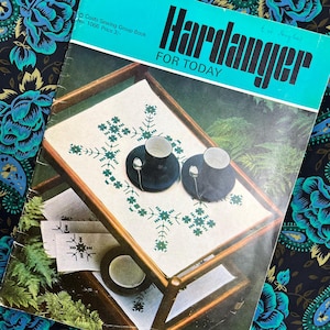 May include: A vintage book titled "Hardanger FOR TODAY" with a teal cover. The image shows a wooden tray with two cups and saucers, and a white cloth with green embroidery. The background features a floral pattern.