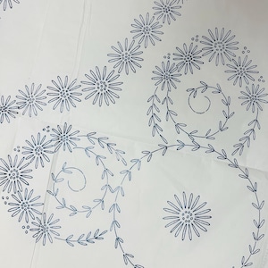 May include: White fabric with a detailed blue floral embroidery design. The embroidery features daisy-like flowers and swirling vines, creating a delicate and intricate pattern. The design is repeated across the fabric.