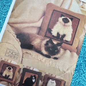 Puede incluir: Un patrón de Vogue Crafts con diseños de cojines con temática de gatos. La imagen muestra un cojín terminado con un diseño de gato siamés, junto con tres diseños de cojines más pequeños: un siamés, un gato blanco y negro y un gato blanco. El patrón es de Linda Carr.