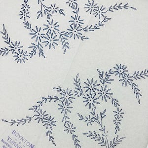Op de afbeelding: Een wit papieren borduurtransfervel met twee donkerblauwe bloemmotieven bestaande uit madeliefachtige bloemen en bebladerde stengels. De tekst "BOYNTON & TURNER'S SUPERIOR TRANSFER BRITISH MAKE" en "10X10 15454" staat in de hoek gedrukt.