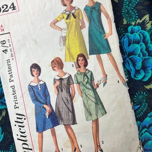 Può includere: Una busta per cartamodello Simplicity Printed Pattern con illustrazioni di cinque diversi modelli di abiti. Gli abiti includono un abito giallo con colletto bianco, un abito verde acqua con fiocco, un abito blu a pois, un abito a quadri e un abito verde a righe.