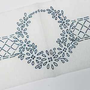 May include: A white fabric transfer featuring a dark blue ink design of a central floral wreath composed of daisy-like flowers. The wreath is flanked on both sides by a geometric diamond lattice pattern, creating a continuous decorative border.