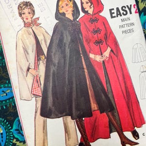Op de afbeelding: Een vintage naaipatroon-envelop met illustraties van drie capes met capuchon in wit, zwart en rood. De tekst op de hoes bevat "EASY", "MAIN PATTERN PIECES" en maataanduidingen variërend van Small tot X Large voor de maten 10 tot 40.