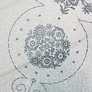 May include: Close-up of a white fabric with a floral embroidery design. The embroidery features a cluster of flowers in the centre, surrounded by a circular pattern and additional floral motifs. The design is in black thread.