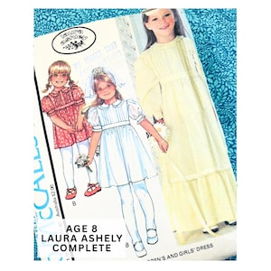 Op de afbeelding: Een McCall's naaipatroon-envelop voor een Laura Ashley kinder- en meisjesjurk, maat 8 jaar. De hoes bevat illustraties van drie jurkvariaties in rood, blauw en geel, met de tekst Laura Ashley en Age 8 duidelijk zichtbaar.
