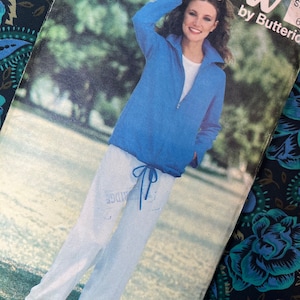 May include: A vintage sewing pattern cover featuring a woman in a blue zip-up top and white sweatpants. The pattern is titled "Easy & Sew" by Butterick. The cover has a worn appearance, suggesting its age.