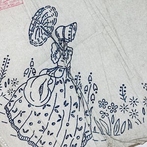 Op de afbeelding: Een wit papieren borduurtransfervel met een donkerblauwe inkttekening van een persoon in een lange jurk en kapje die een parasol vasthoudt, omringd door bloemmotieven. De tekst "Leach's" en "MADE IN ENGLAND" is in het rood gedrukt.