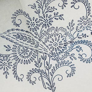 Könnte beinhalten: Eine weiße Stoffoberfläche mit einem dunkelblauen Tintentransfermuster eines Blumenstraußes. Das Design besteht aus stilisierten Blumen, Blättern und wirbelnden Rankenelementen mit feinen Liniendetails, die eine dekorative botanische Illustration bilden.
