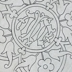 Pode incluir: Uma folha de tecido cinza claro com um padrão de bordado de linhas pretas. O design consiste num motivo circular central contendo uma letra de monograma ornamentada, rodeado por um arranjo simétrico de flores e folhas estilizadas.
