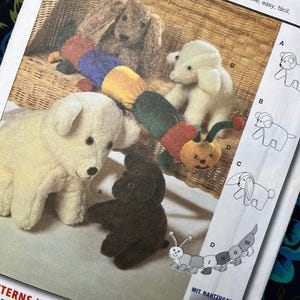 May include: A sewing pattern envelope featuring illustrations and photographs of four stuffed animal toys: a large cream dog, a small dark brown dog, a cream puppy, and a multi-coloured segmented caterpillar with red, blue, yellow, and green sections.