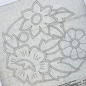 Puede incluir: Una hoja de papel transfer blanco translúcido con un dibujo de líneas negras de un arreglo floral con tres tipos de flores distintos y varias hojas. La esquina inferior incluye texto impreso y un icono de tijeras para referencia.