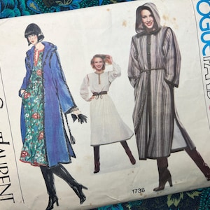 Op de afbeelding: Een Vogue Paris Original naaipatroon-envelop, nummer 1738, van Yves Saint Laurent. De illustratie toont drie kledingontwerpen: een blauwe jas met capuchon over een bloemenjurk, een witte jurk met een bruine riem en een gestreepte jas met capuchon.