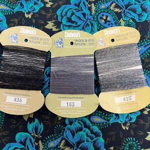 May include: Three spools of Chadwick's thread in shades of black, grey, and white. Each spool is labelled with the text "WOOL & 85% NYLON 15%" and a product number. The background is a dark fabric with a floral pattern.