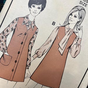 Op de afbeelding: Een naaipatroon-envelop met illustraties van twee jurkstijlen. Stijl A toont een doorknoopjurk met een bloemrijke blouse met lange mouwen. Stijl B toont een mouwloze jurk met een blouse met strikkraag. Tekst bevat B, A en A&B.