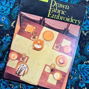 Peut inclure: Un livre de couture vintage intitulé "Drawn Fabric Embroidery" est posé sur une illustration de table. Le livre est noir avec du texte jaune. La table comprend des assiettes, des verres et des couverts, avec une nappe crème et une table marron.