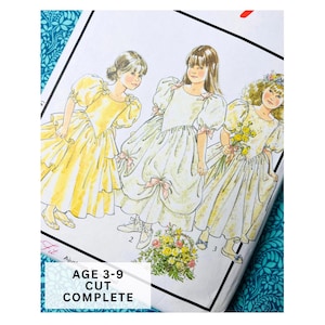 Op de afbeelding: Een vintage naaipatroon met illustraties van drie meisjes in jurken. De jurken zijn geel en wit, met strikken en bloemendetails. De tekst op het patroon luidt "AGE 3-9 CUT COMPLETE."