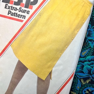 Op de afbeelding: Een Simplicity E.S.P. Extra-Sure patroon envelop met een gele knielange rok met een knoopsluiting aan de zijkant. De witte envelop bevat rode grafische lijnen en tekst, en toont een gele rok met een rechte pasvorm en knopen.