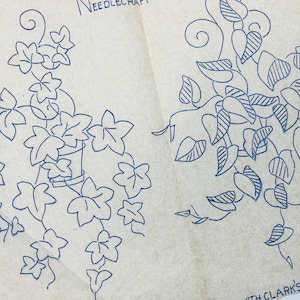 May include: A vintage embroidery transfer pattern on off-white paper featuring two blue ink illustrations of trailing ivy plants in pots. The text NEEDLECRAFT and WITH CLARK'S ANCHOR are visible in blue ink on the paper surface.