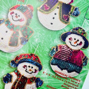 Könnte beinhalten: Set mit vier skurrilen Schneemann-Ornamenten, jeweils ca. 12,7 cm x 15,2 cm groß. Die Schneemänner sind weiß mit bunten Paillettenhüten, Schals und Jacken. Entworfen von Bonnie Smith.